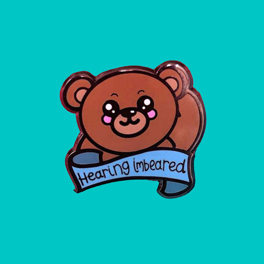 Hearing Imbeared Enamel Pin - Hearing Impaired on a blue background. The enamel pin has an adorable brown teddy bear with big sparkly eyes and rosy cheeks. The bear is holding one hand to it's ear. There is a blue banner under the bear with hearing imbeared written across it in black writing. The enamel pin is designed to raise awareness for hearing impairments