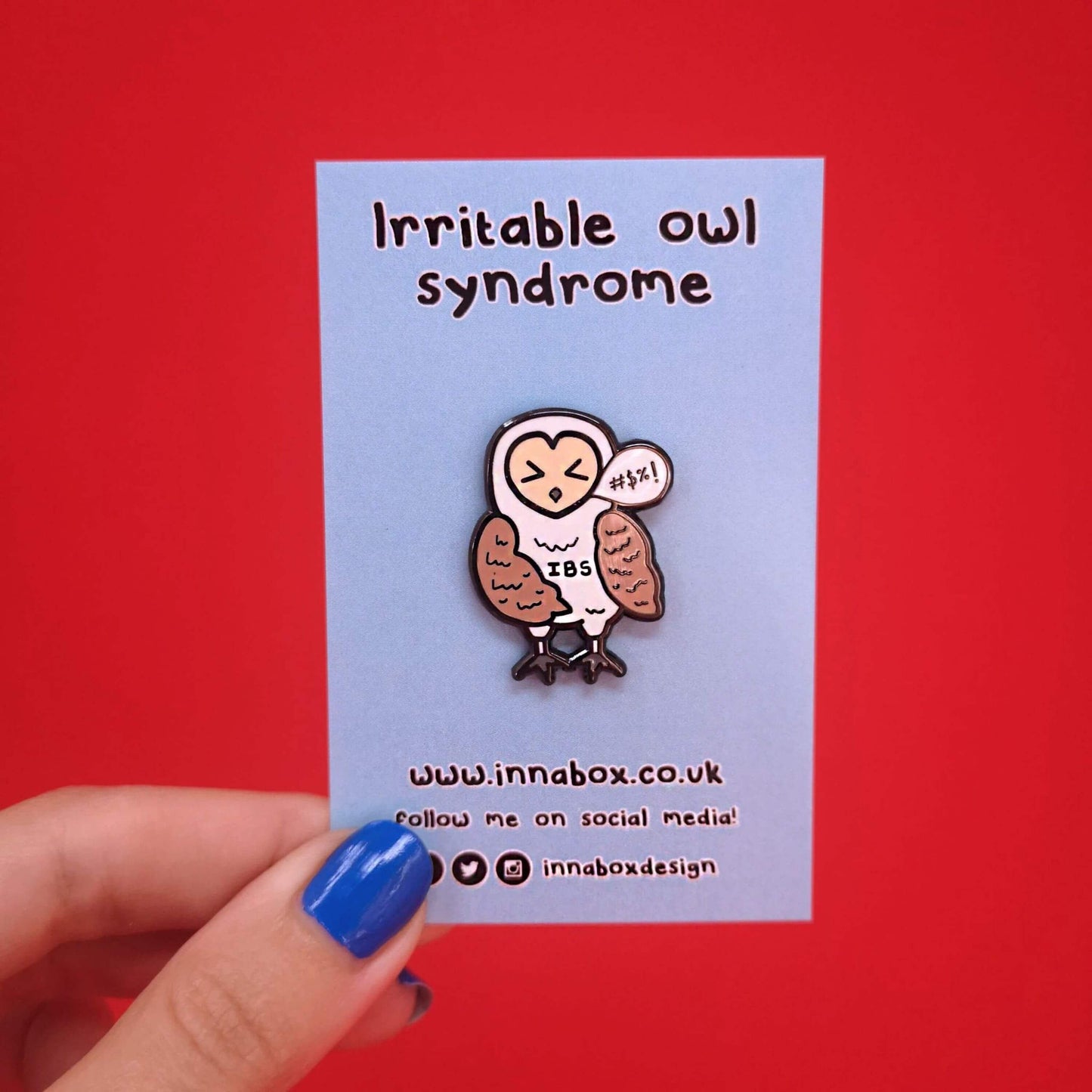 Irritable Owl Syndrome Enamel Pin - Irritable bowel syndrome (IBS) on backing card in front of a red background. The enamel pin is of a barn owl with a sweary text speech bubble and has its eyes shut with the letters IBS written on its belly. Hand drawn design to raise awareness for Irritable bowel syndrome (IBS)