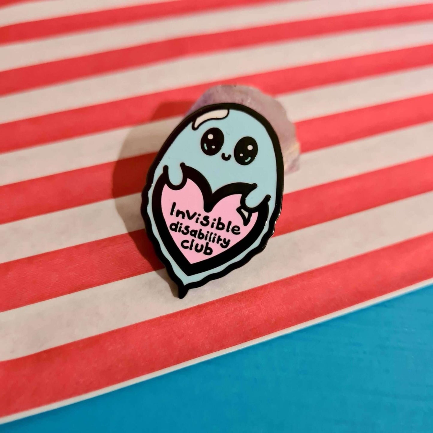 Invisible Disability Club Enamel Pin shown on a red and white striped paper bag on a blue background. The enamel pin is of a cute smiling ghost holding a pink heart with text saying invisible disability club. The enamel pin is designed to raise awareness for hidden and chronic disabilities