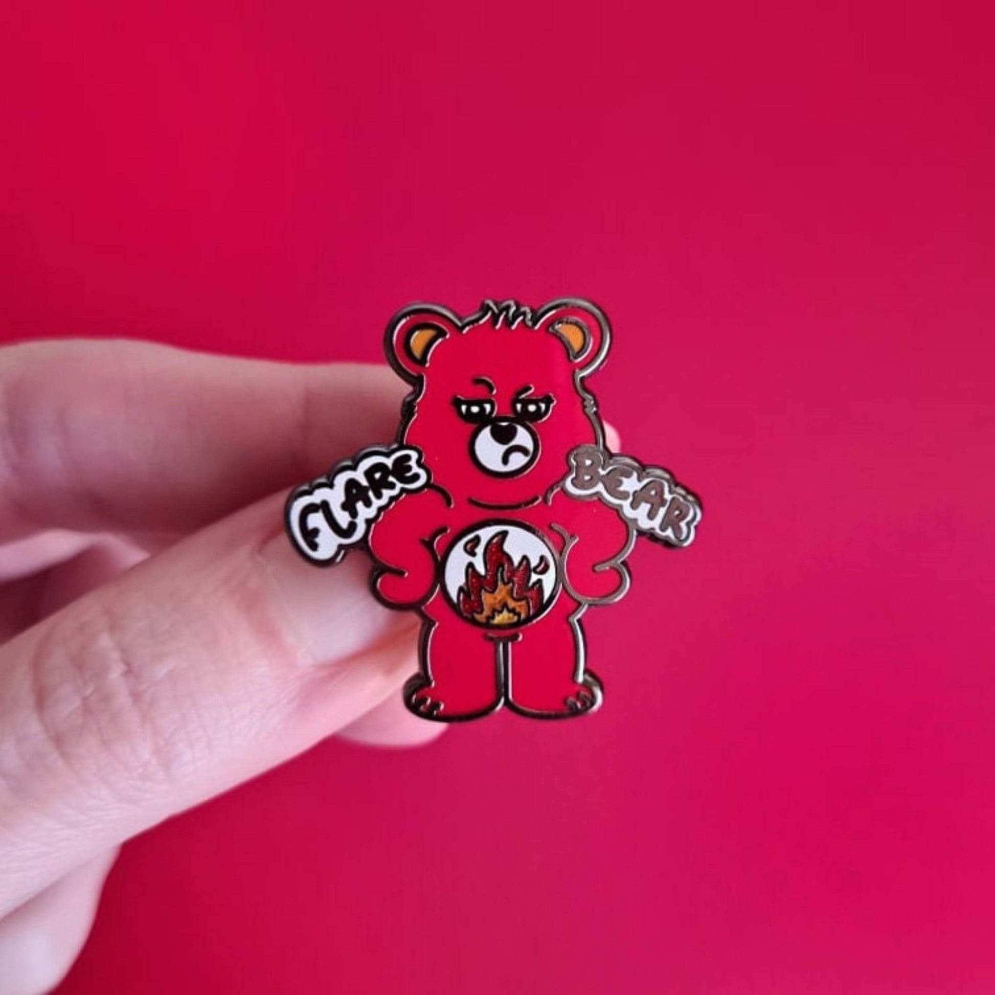 Flare Bear enamel pin shown held by a hand in front of a red background. The enamel pin is of a red bear with a fed up expression and hands on its hips. There is a white circle on its belly with flames inside. Flare Bear is written on the pin. The enamel pin is designed to raise awareness for chronic illness flare ups.