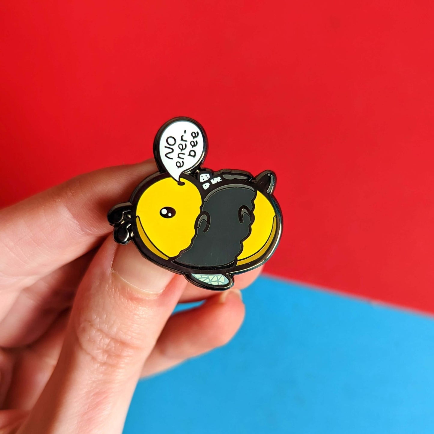 Low Energy Bee Enamel Pin held in front of a red and blue background. The enamel pin is a cute yellow and black bee lying on it's wings with its arms, legs, little belly and stinger in the air. The bee has a speech bubble coming from it's mouth with 'NO ener-bee' written inside in black writing. Hand drawn design made to raise awareness for chronic fatigue.