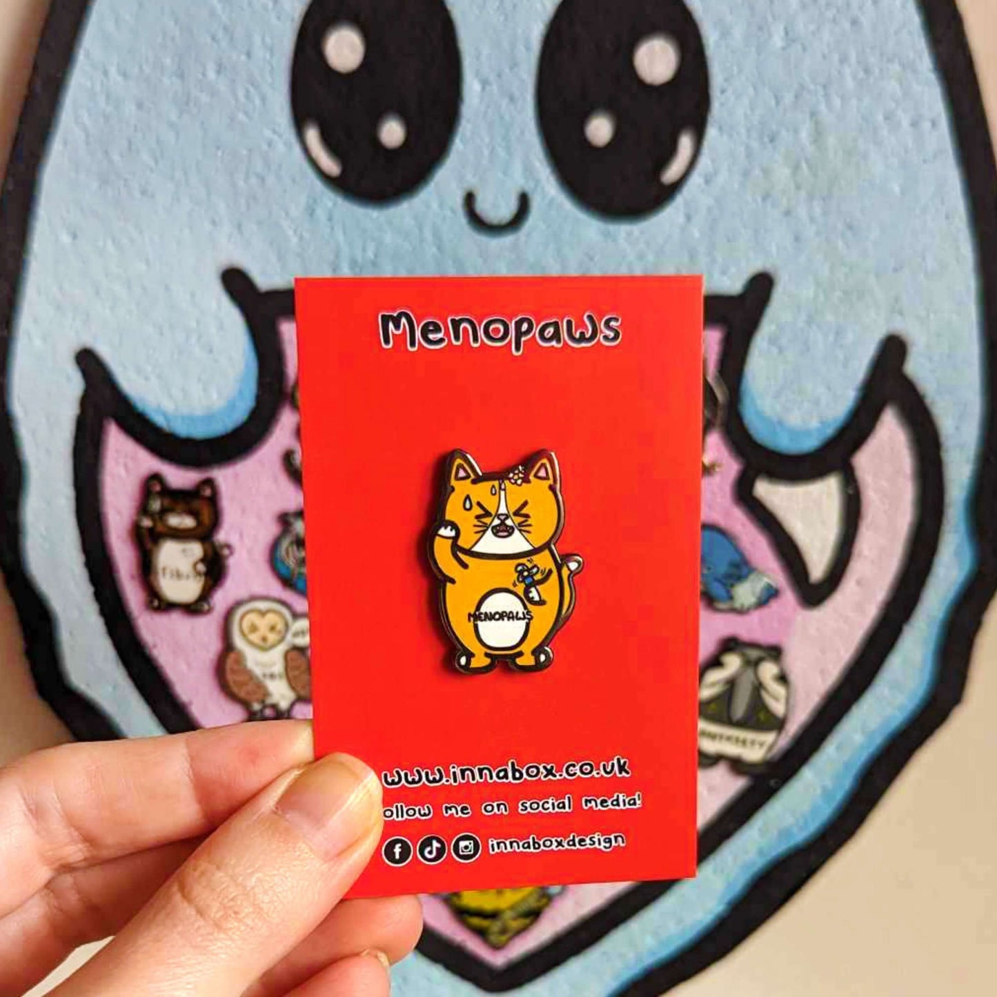 Menopaws Enamel Pin - Menopause shown on red backing card held in front of a cork board with various enamel pins on. The enamel pin is of a menopausal ginger and white cat with a daisy in it's hair. The cat is holding an electric fan and is looking hot with sweat droplets on it's face. Hand drawn design made to raise awareness for menopause