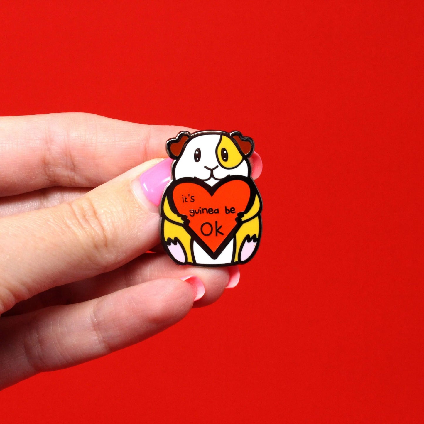 Guinea Pig Mental Health Enamel Pin shown held in front of a red background. The enamel pin is of a smiling guinea pig holding a big red heart with the text it's guinea be ok inside. The enamel pin is deigned to raise awareness for mental health.