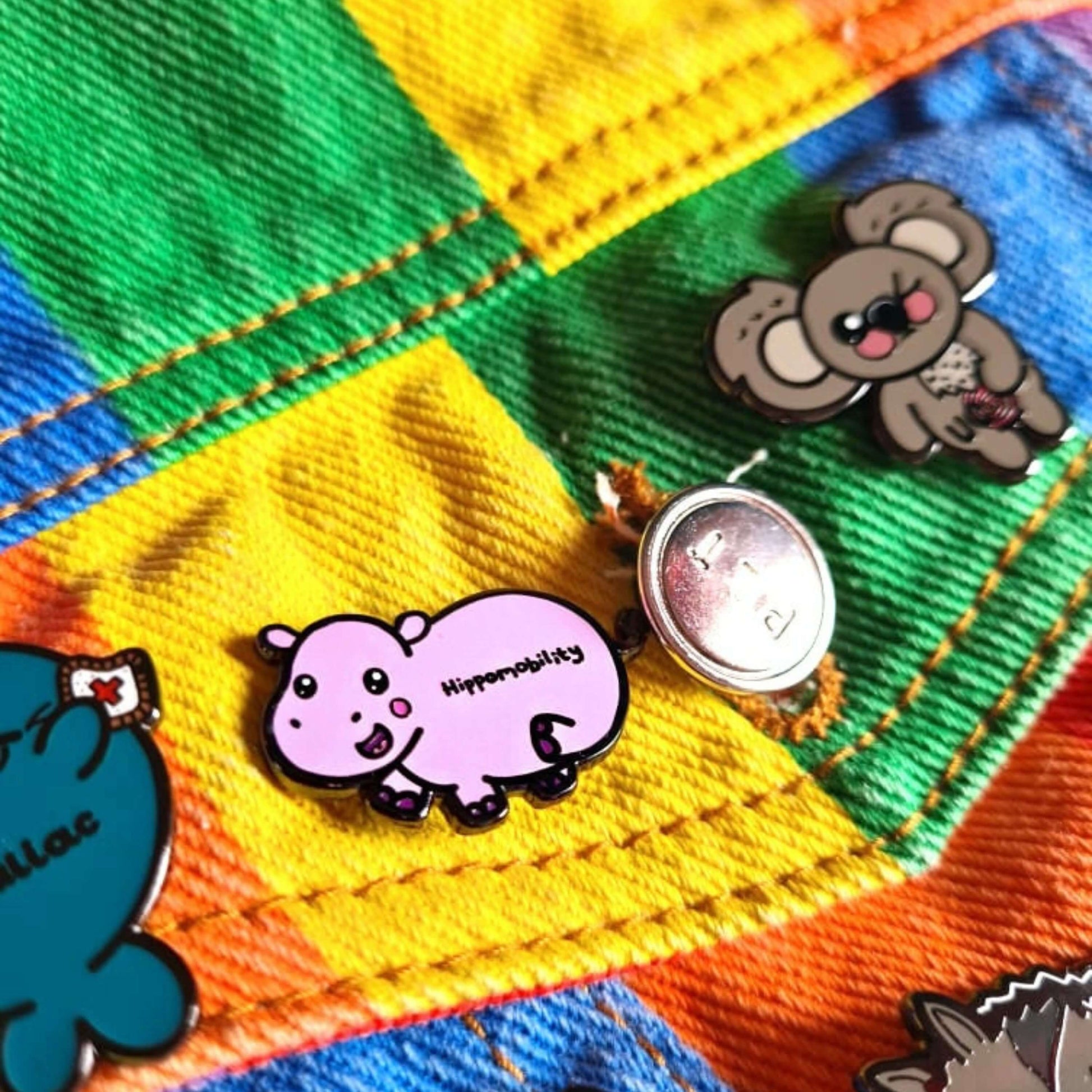 Hippmobility Enamel Pin - Hyper Mobility pinned on a rainbow denim jacket with other innabox enamel pins. The enamel pin is of a pink happy hippo with the text hippomobility written on its stomach. The enamel pin is designed to raise awareness for hyper mobility