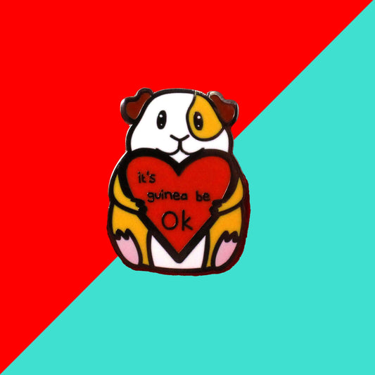 Guinea Pig Mental Health Enamel Pin shown on a red and blue background. The enamel pin is of a smiling guinea pig holding a big red heart with the text it's guinea be ok inside. The enamel pin is deigned to raise awareness for mental health.