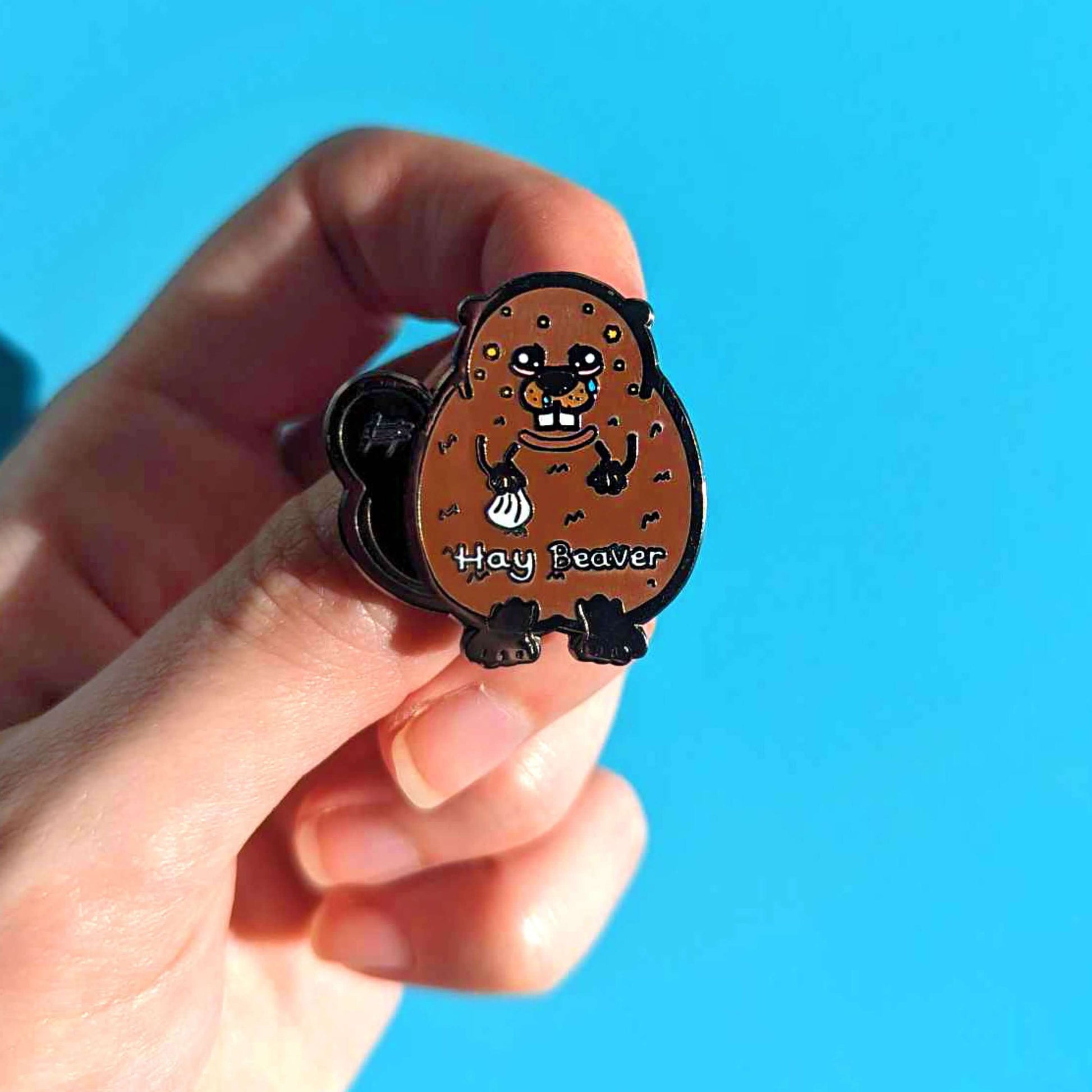 Hay Beaver Enamel Pin - Hay Fever held in front of a blue background. The enamel pin is a brown beaver with watery eyes, dripping nose and yellow spots on face and is holding a tissue with it's little hand. Hay beaver is written across its belly. The enamel pin is designed to raise awareness for hay fever or allergic rhinitis