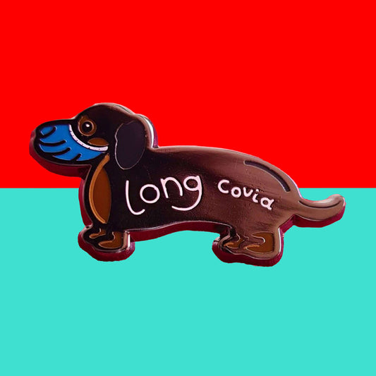 Long Covid Enamel Pin shown on red and blue background. The enamel pin is of a brown and black sausage dog wearing a blue medical mask over mouth and nose and 'long covid' written across it's body in white. Hand drawn design made to raise awareness for long covid.