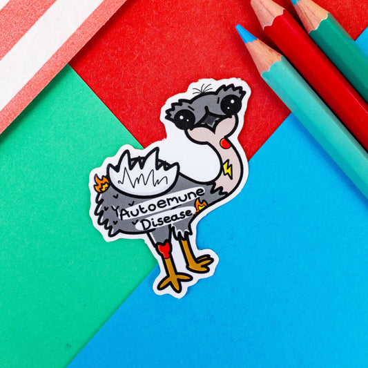 Autoemune Sticker - Autoimmune Disease shown on a blue, red and green background with colouring pencils and a red stripe candy bag. The sticker is a grey and white emu bird with various problems highlighted with lightning bolts, flames and red circles with the words autoemune disease written across its belly. Design created to raise awareness for autoimmune diseases.