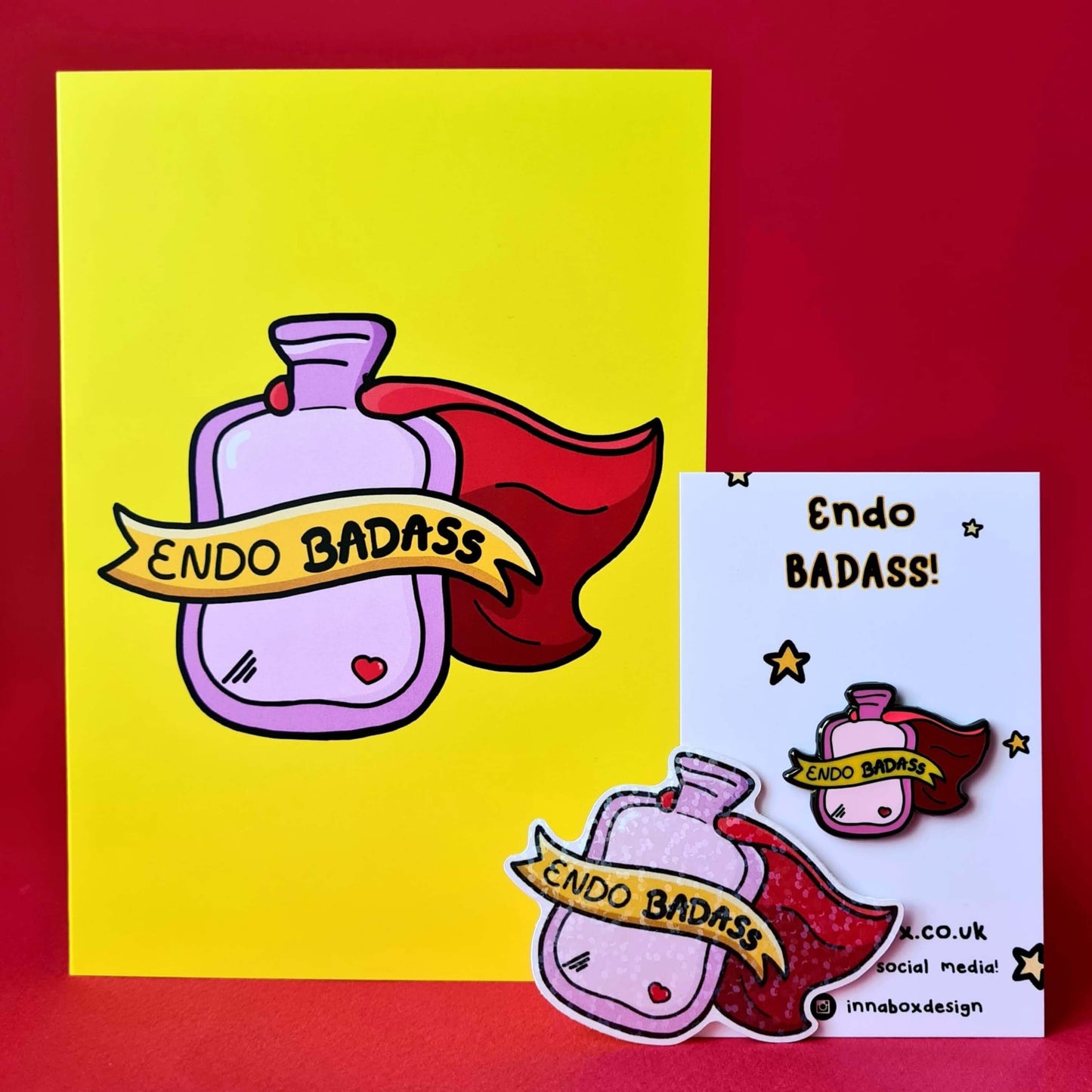 The Endo Badass Card - Endometriosis on a red background with other endo themed innabox products, a pin badge and holographic sticker. The yellow based card features a pink hot waterbottle with a small red heart, a red cape and a yellow banner across reading 'endo badass'. Hand drawn design is raising awareness for Endometriosis and pelvic pain.