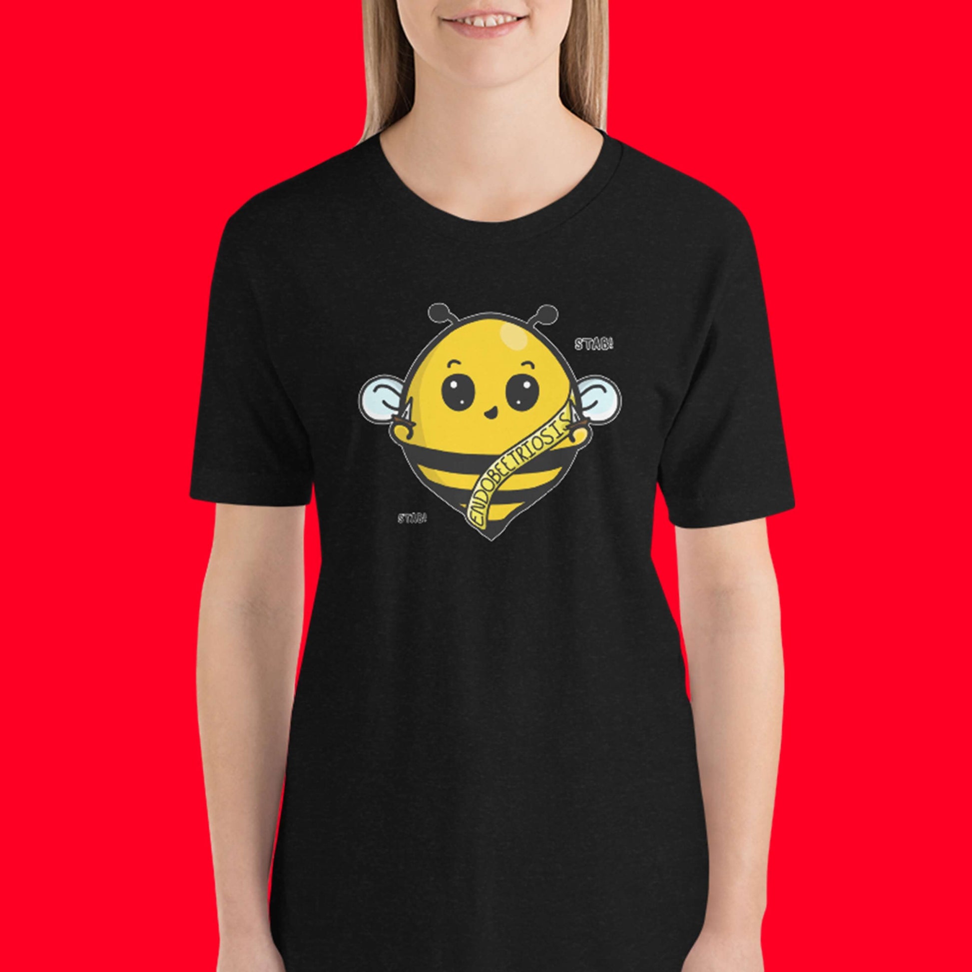The Endobeetriosis Tee - Endometriosis being modelled by a femme person facing forward smiling on a red background. The black short sleeve tshirt features a smiling happy bumblebee holding small daggers with a yellow banner reading 'endobeetriosis' with the words 'stab! stab!' surrounding the buzzy bee. The hand drawn design is raising awareness for endometriosis.