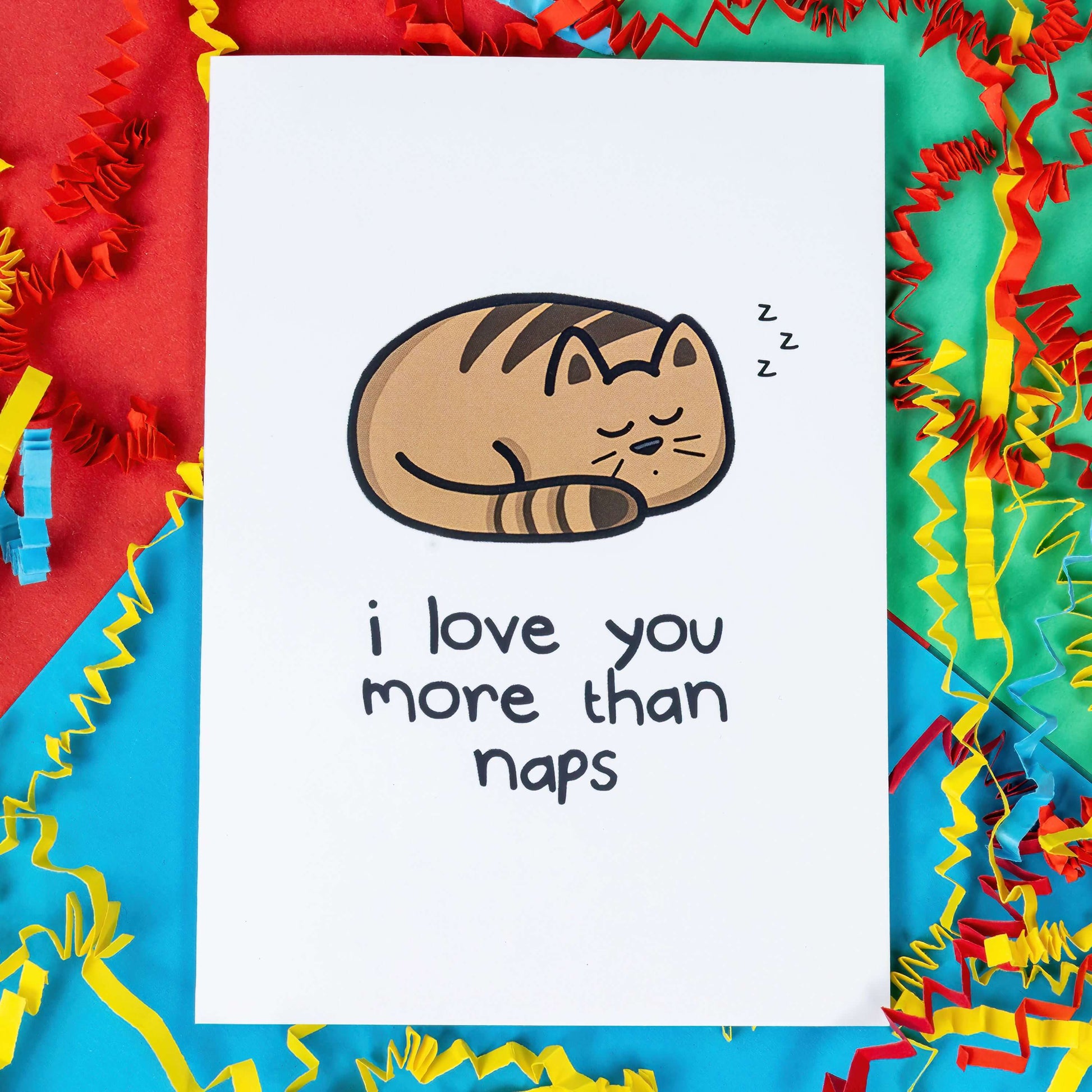 I love You More Than Naps - Cat Card on a red, blue and green background with coloured card confetti. The white card has an illustration of a sleeping tabby cat with black text underneath that reads 'I love you more than naps'.