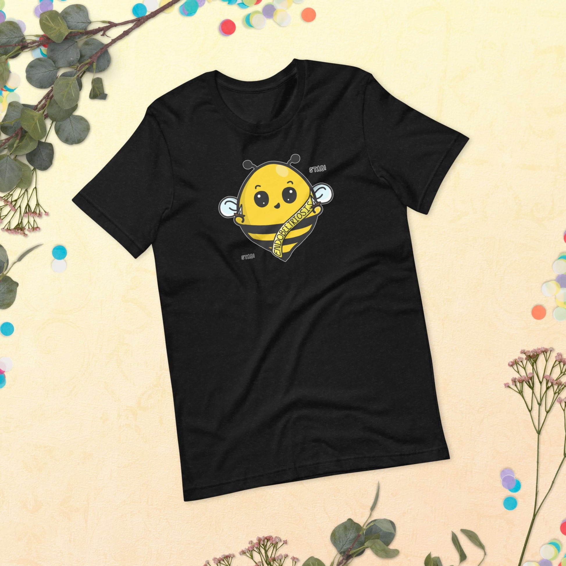 The Endobeetriosis Tee - Endometriosis laying on a pastel yellow background with multicoloured confetti and plant foliage. The black short sleeve tshirt features a smiling happy bumblebee holding small daggers with a yellow banner reading 'endobeetriosis' with the words 'stab! stab!' surrounding the buzzy bee. The hand drawn design is raising awareness for endometriosis.