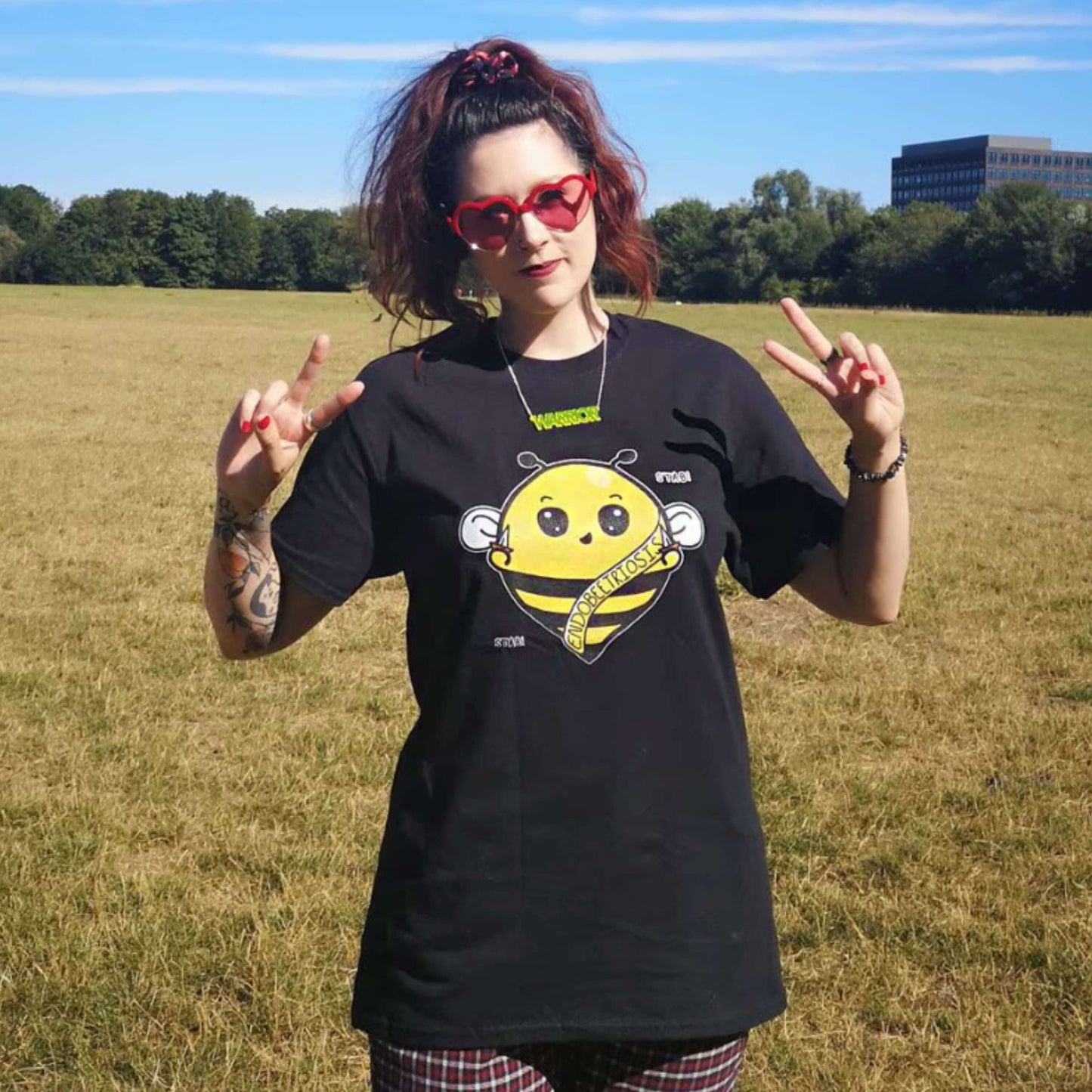 The Endobeetriosis Tee - Endometriosis being modelled by Nikky outside in a grass field wearing red heart sunglasses and red checkered trousers, she is smiling with both hands up in a peace sign. The black short sleeve tshirt features a smiling happy bumblebee holding small daggers with a yellow banner reading 'endobeetriosis' with the words 'stab! stab!' surrounding the buzzy bee. The hand drawn design is raising awareness for endometriosis.
