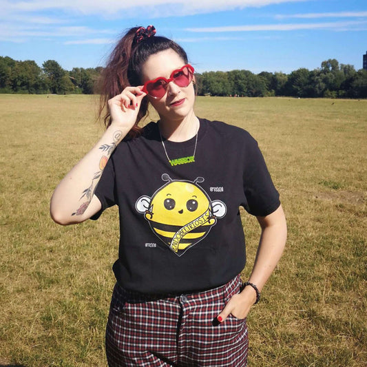 The Endobeetriosis Tee - Endometriosis being modelled by Nikky outside in a grass field wearing red heart sunglasses and red checkered trousers, she is smiling with one hand in her pocket and the other holding her glasses. The black short sleeve tshirt features a smiling happy bumblebee holding small daggers with a yellow banner reading 'endobeetriosis' with the words 'stab! stab!' surrounding the buzzy bee. The hand drawn design is raising awareness for endometriosis.