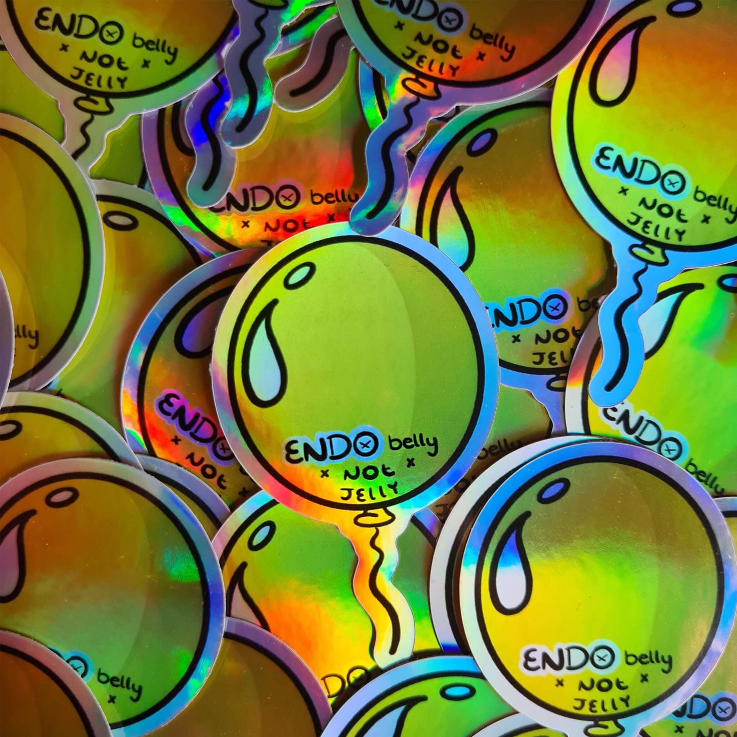 The Endo Belly Sticker on a pile of multiple copies of the sticker. The yellow balloon shape sticker has bottom black text reading 'ENDO belly not jelly'. The hand drawn design is raising awareness for endometriosis.