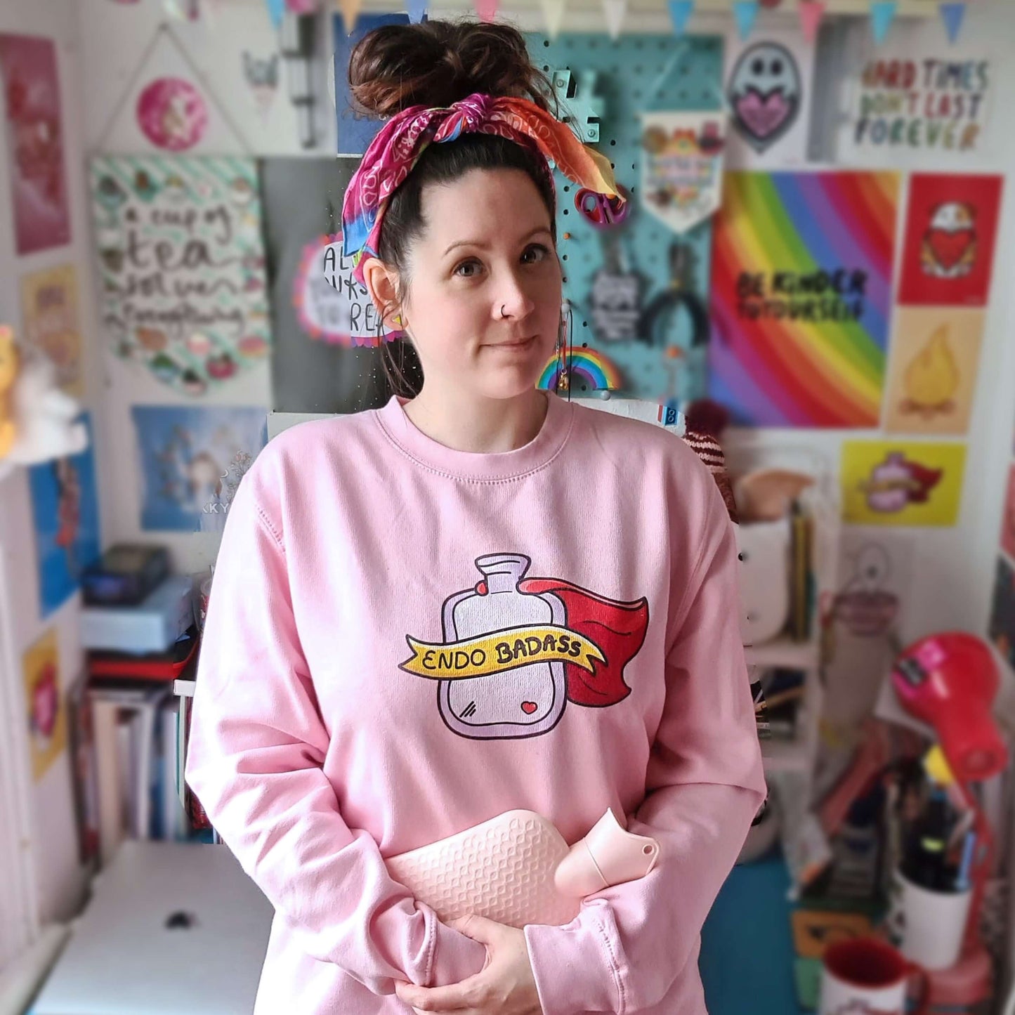 The Endo Badass Sweater - Endometriosis being modelled by Nikky with brown hair in a messy bun smiling clutching a pink hot waterbottle in front of a wall of their artwork. A pastel bubblegum baby pink sweatshirt jumper with a pink hot waterbottle with a small red heart, red cape and yellow banner reading 'endo badass' in black text. The hand drawn design is raising awareness for endometriosis and pelvic pain.
