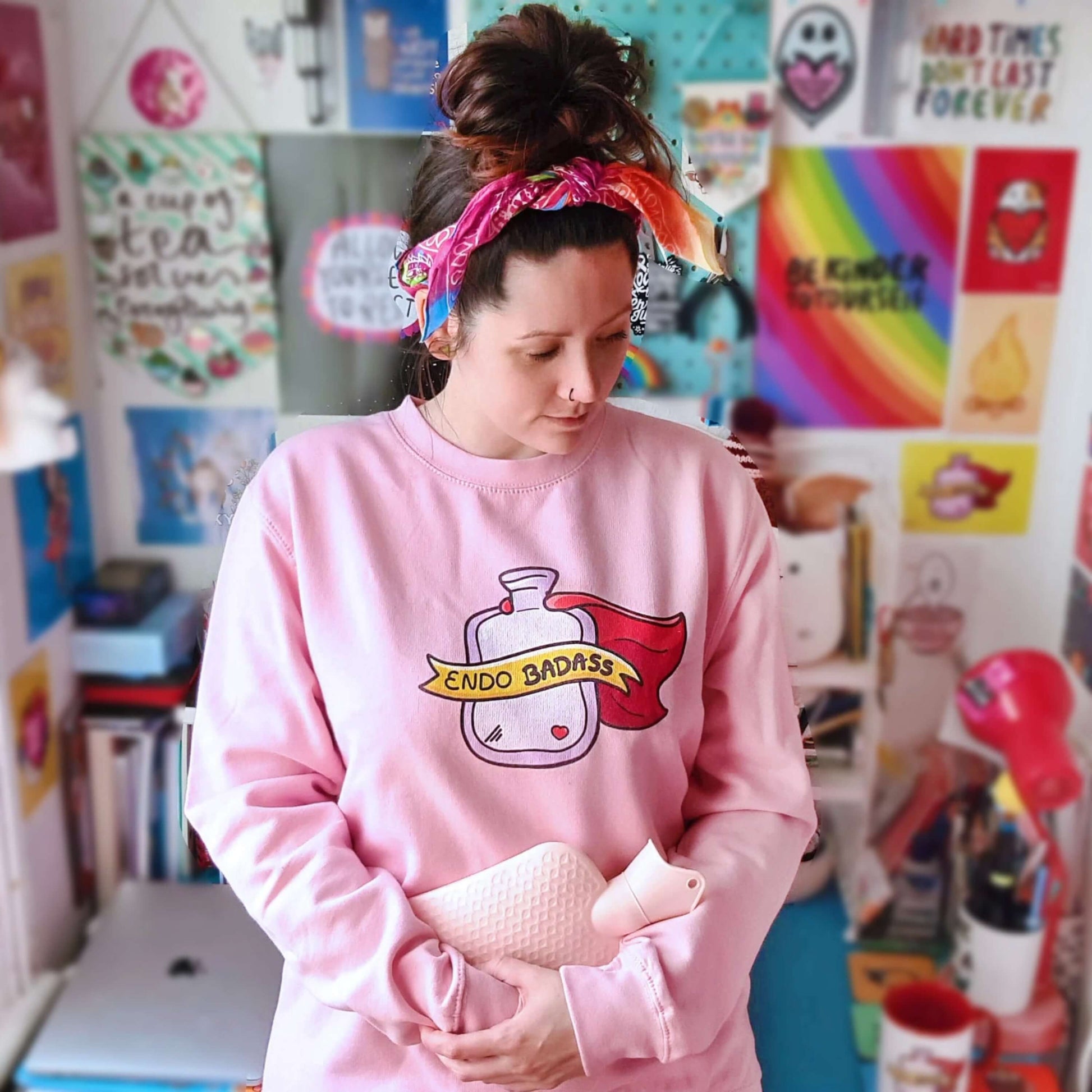 The Endo Badass Sweater - Endometriosis being modelled by Nikky with brown hair in a messy bun looking down to the right clutching a pink hot waterbottle in front of a wall of their artwork. A pastel bubblegum baby pink sweatshirt jumper with a pink hot waterbottle with a small red heart, red cape and yellow banner reading 'endo badass' in black text. The hand drawn design is raising awareness for endometriosis and pelvic pain.