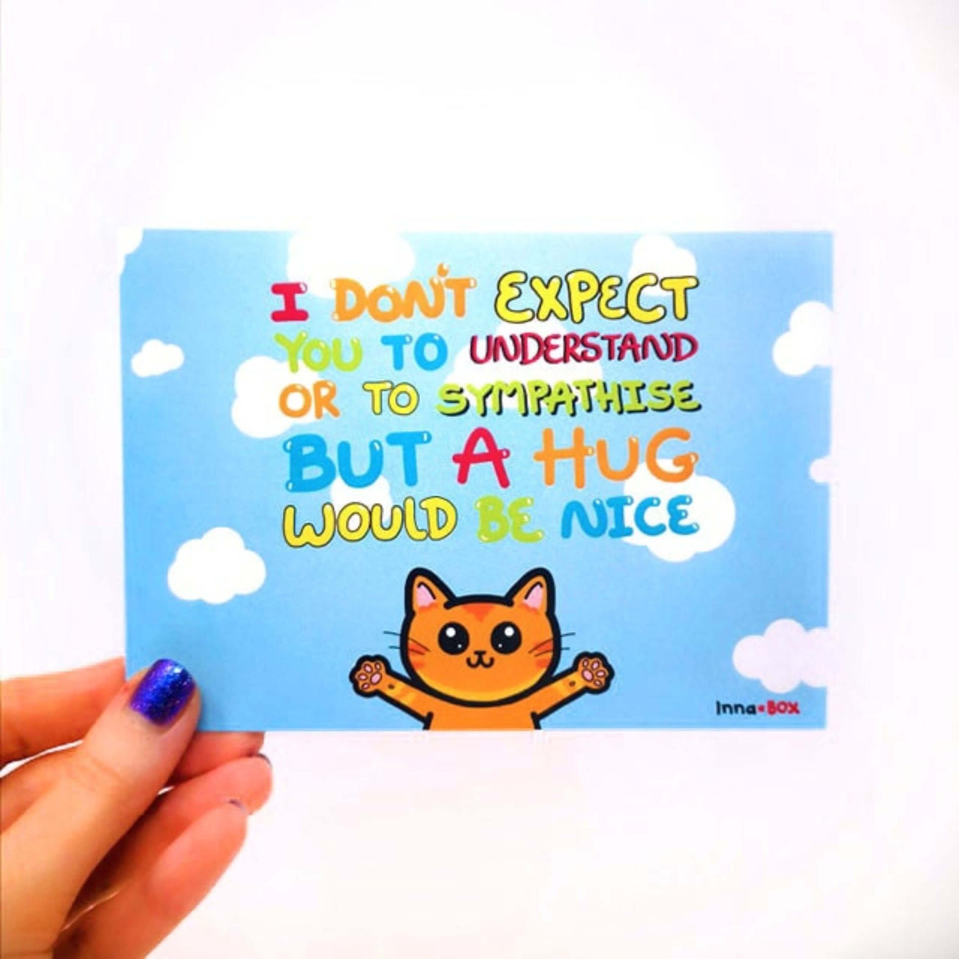 A Hug Would Be Nice - Cat Postcard held in front of a white background. The light blue postcard has rainbow bubble text saying 'I don't expect you to understand or to sympathise but a hug would be nice'. Underneath the writing is an illustration of a ginger cat with big eyes and smile with it's arms wide open. The hand drawn design is made to raise awareness for invisible and chronic illnesses.