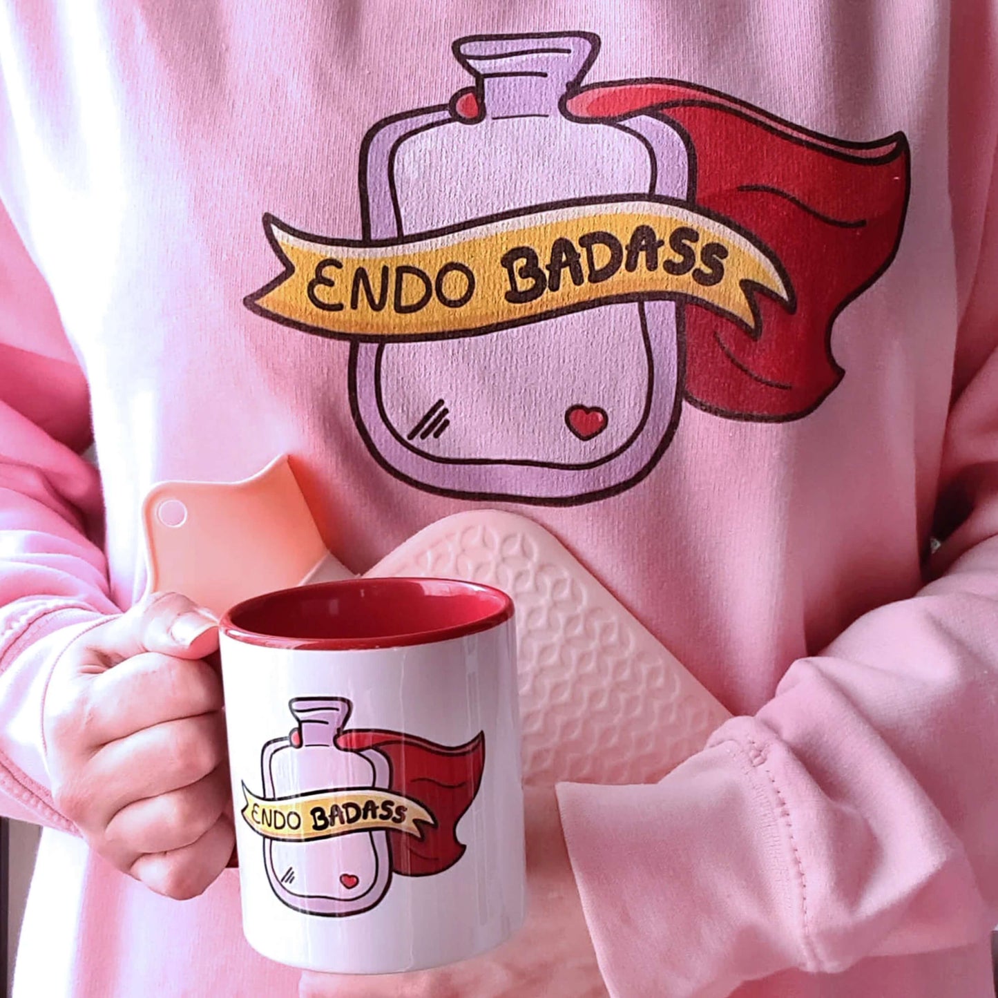 The Endo Badass Sweater - Endometriosis being modelled by Nikky clutching a pink hot waterbottle and matching innabox endometriosis mug. A pastel bubblegum baby pink sweatshirt jumper with a pink hot waterbottle with a small red heart, red cape and yellow banner reading 'endo badass' in black text. The hand drawn design is raising awareness for endometriosis and pelvic pain.