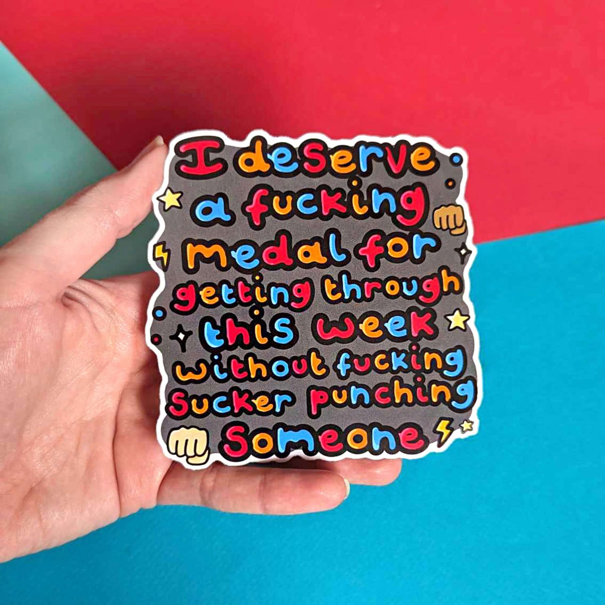 I Deserve A Medal Sticker of the words I deserve a fucking medal for getting through this week without fucking sucker punching someone' in bubble text with hand, lightning bolts, sparkles and star emojis. It is held in a hand in front of a red, blue and green background.