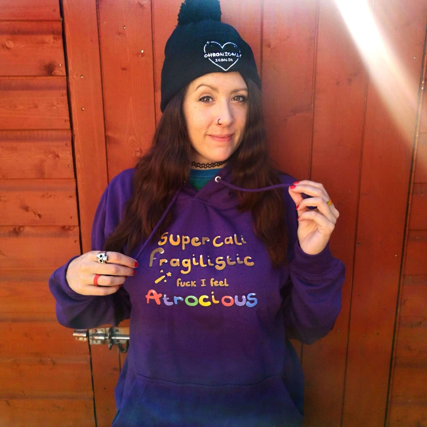 I Feel Atrocious Hoodie worn by Nikky the owner . The plum coloured hoodie has gold text on it that says 'supercali fragilistic f*** I feel' and then 'atrocious' in rainbow coloured text. Design to raise awareness for chronic illness