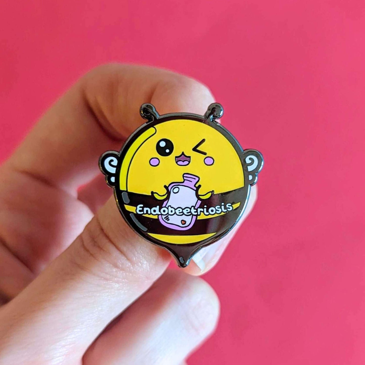 Endobeetriosis 2.0 Bee Enamel Pin - Endometriosis held over a red background. The enamel pin is a bumble bee with a smiley face and holding a pink hot watterbottle in its hands, across its middle is white text that reads endobeetriosis. The enamel pin is designed to raise awareness for endometriosis