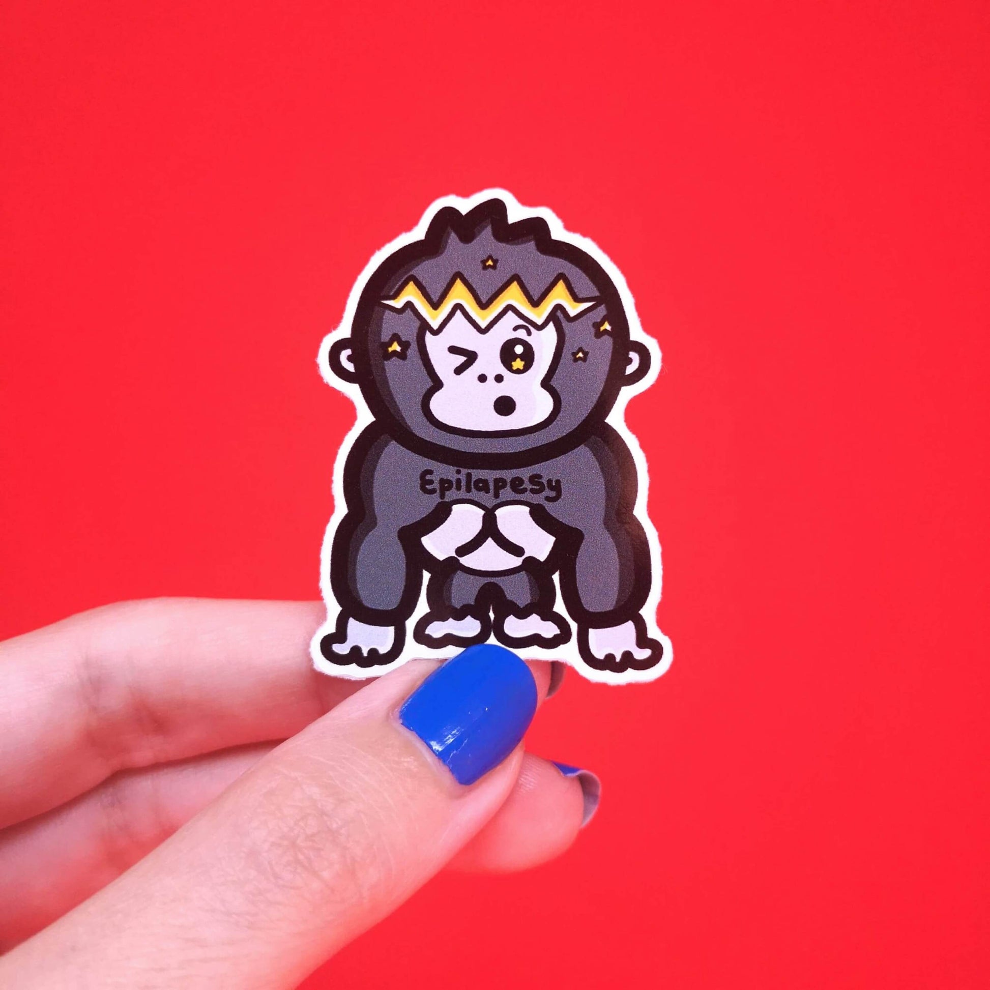 The Epilapesy - Epilepsy Ape Sticker being held over a red background. A gorilla ape shape sticker with a shocked open mouth expression with a yellow lightning bolt and stars across its head, across its chest in black reads 'epilapesy'. The hand drawn design is raising awareness for epilepsy and seizures.