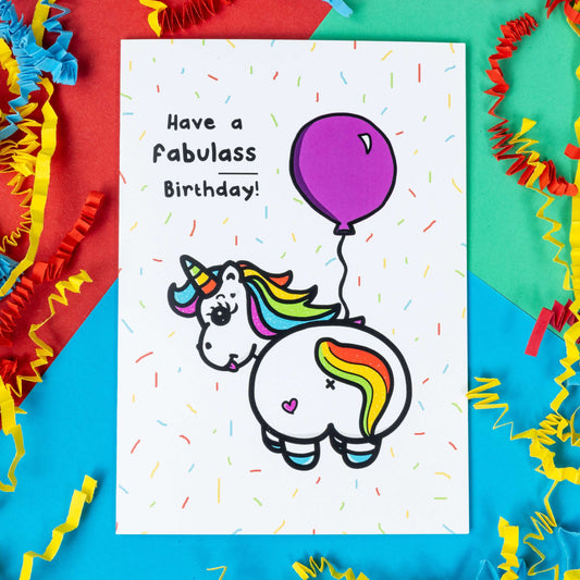 a white greeting card with an illustration of a unicorn with it's bum to the camera and looking back over it's shoulder with it's tongue out. The unicorn has rainbow coloured hair and horn and a pink love heart on it's bum and is holding a pink balloon. There is rainbow confetti all over the card and 'Have a fabulass Birthday!' is written in black with 'ass' underlined. The background of the photo is red, green and blue card with red and yellow paper decorations.
