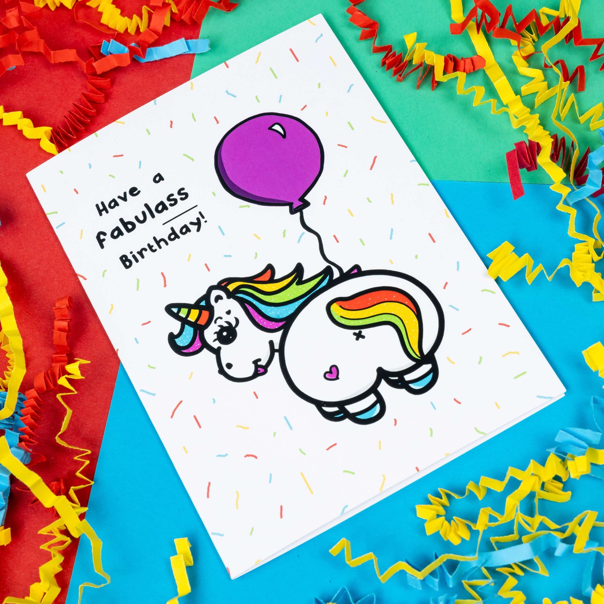 a white greeting card with an illustration of a unicorn with it's bum to the camera and looking back over it's shoulder with it's tongue out. The unicorn has rainbow coloured hair and horn and a pink love heart on it's bum and is holding a pink balloon. There is rainbow confetti all over the card and 'Have a fabulass Birthday!' is written in black with 'ass' underlined. The background of the photo is red, green and blue card with red and yellow paper decorations.