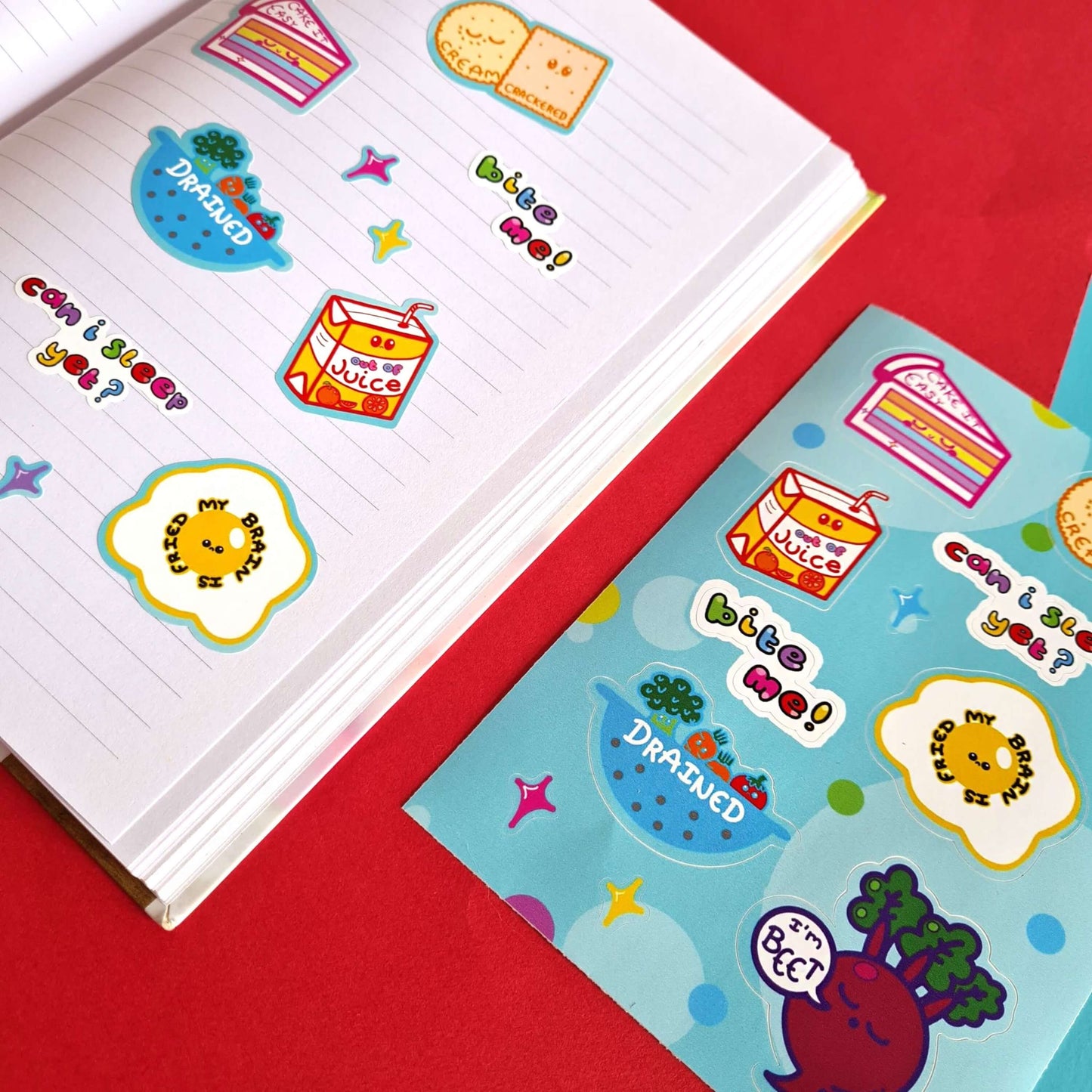 The Fatigued Foods Sticker Sheet - A6 Sticker Sheet on a red and blue background. Sticker sheet of a smiling rainbow cake with text reading 'cake it easy', two snoozing cream crackers with text reading 'cream crackered', a sad fruit juice box with text 'out of juice', a sad fried egg with text 'my brain is fried', a sieve filled of sad vegetables with the text 'drained', a sleeping beetroot with a speech bubble reading 'I'm beet' as well as rainbow text reading 'can I sleep yet?' and 'bite me'.