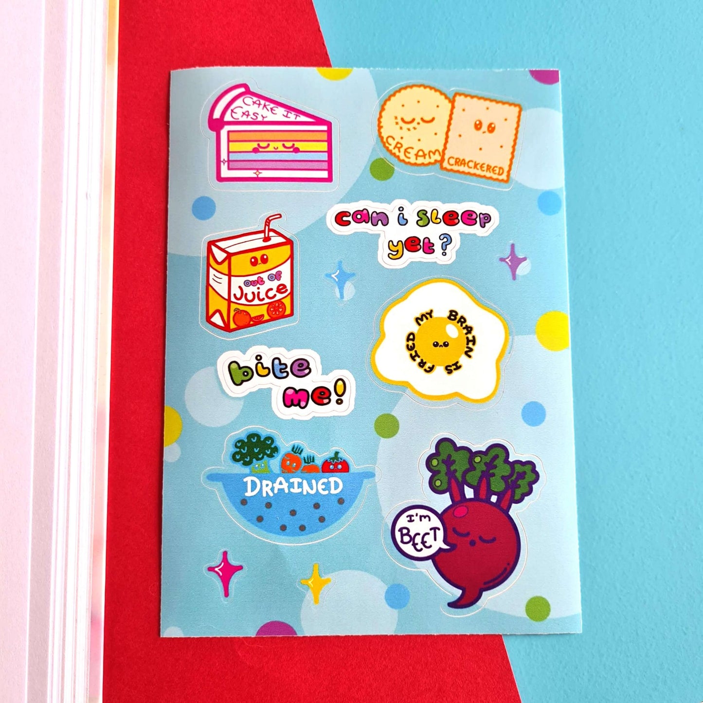 The Fatigued Foods Sticker Sheet - A6 Sticker Sheet on a red and blue background. Sticker sheet of a smiling rainbow cake with text reading 'cake it easy', two snoozing cream crackers with text reading 'cream crackered', a sad fruit juice box with text 'out of juice', a sad fried egg with text 'my brain is fried', a sieve filled of sad vegetables with the text 'drained', a sleeping beetroot with a speech bubble reading 'I'm beet' as well as rainbow text reading 'can I sleep yet?' and 'bite me'.