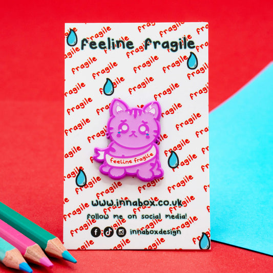Front-facing shot of the Feeline Fragile Cat Enamel Pin on its Innabox backing card. The purple enamel cat features tearful eyes and a “feeline fragile” banner, photographed on red and blue card.