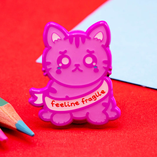 Close-up photo of Innabox’s Feeline Fragile Cat Enamel Pin featuring a cute purple pink crying cat with the words “feeline fragile” on a pink banner. Shown on a bright red background with colourful pencils.