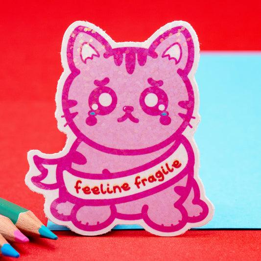 Cute pink glitter cat sticker with teary eyes and the phrase "feeline fragile" on a red and blue background. Created by Innabox, a small UK brand supporting chronic illness and hidden disability awareness.