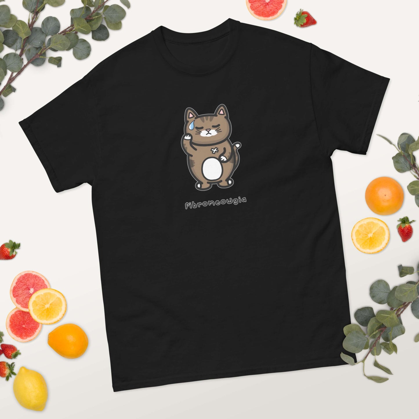 The Fibromeowgia Cat Tee - Fibromyalgia Syndrome laying on a white background with fruit slices and green foliage. The black short sleeve tshirt features a sad brown tabby cat with a sweat droplet clutching its body with a white bandaid and bottom text reading 'fibromeowgia'. The hand drawn design is raising awareness for fibro fibromyalgia syndrome.