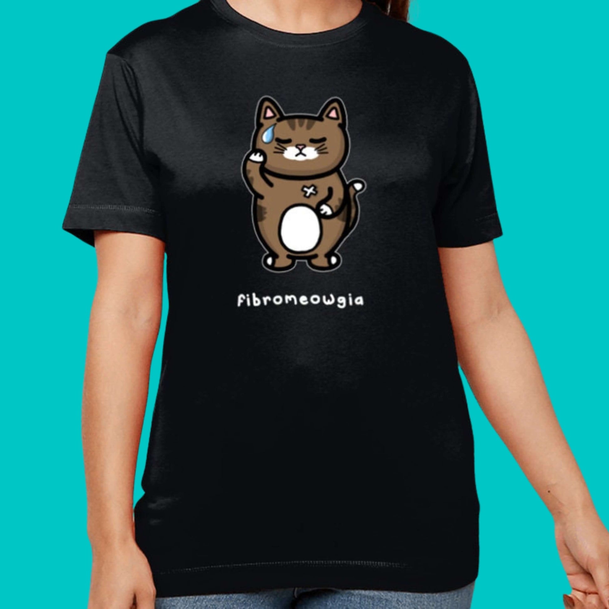 The Fibromeowgia Cat Tee - Fibromyalgia Syndrome modelled by a femme person with brown hair on a blue background. The black short sleeve tshirt features a sad brown tabby cat with a sweat droplet clutching its body with a white bandaid and bottom text reading 'fibromeowgia'. The hand drawn design is raising awareness for fibro fibromyalgia syndrome.