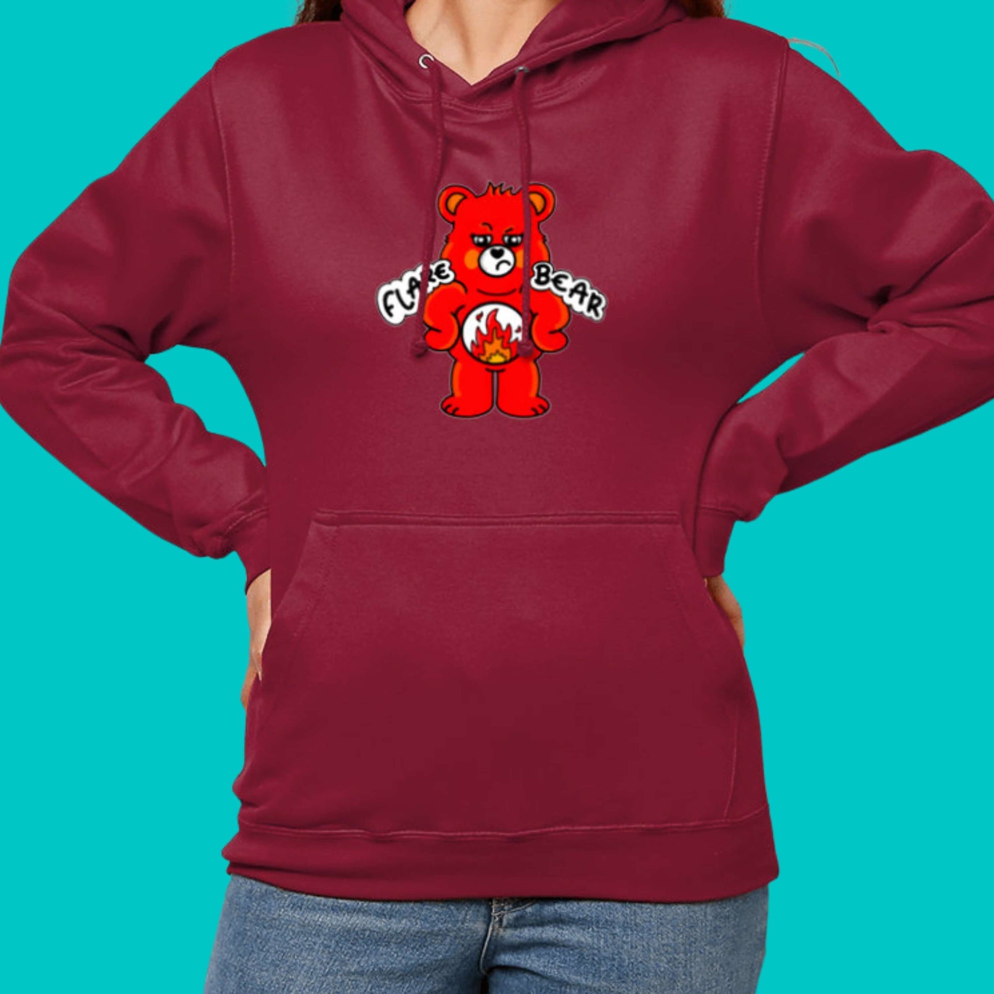 Flare Bear hoodie jumper in red hot chilli modelled by a femme person with brown hair wearing blue jeans on a blue background. The hoodie is of a red bear with a fed up expression and hands on its hips. There is a white circle on its belly with flames inside. Flare Bear is written on the hoodie. The hoodie is designed to raise awareness for chronic illness flare ups.