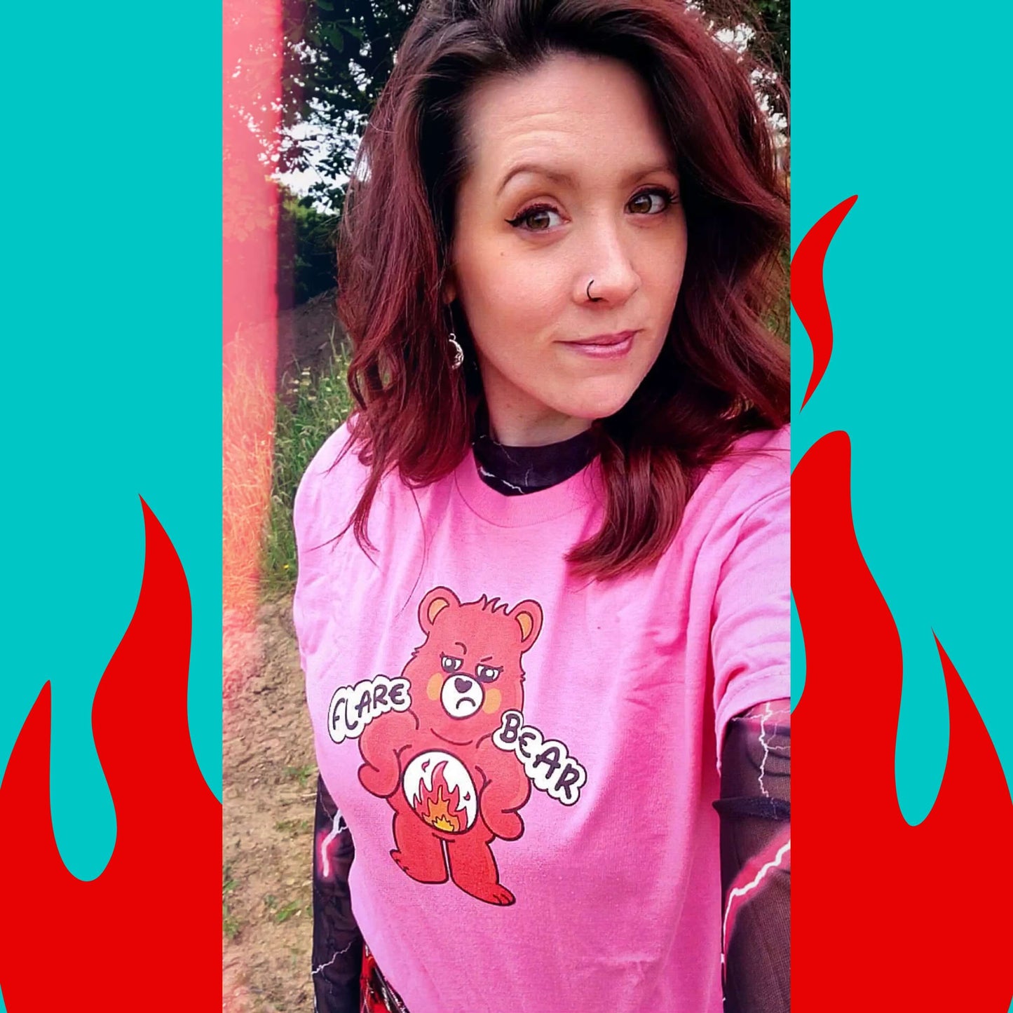 Nikky wearing a pink Flare Bear T-shirt stood outside with red tartan trousers, a pink and black lightning bolt mesh top. The pastel pink tee is of a red bear with a fed up expression and hands on its hips. There is a white circle on its belly with flames inside. Flare Bear is written on the middle. The tshirt is designed to raise awareness for chronic illness flare ups.