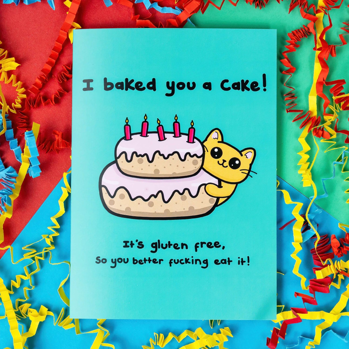 A turquoise blue card that has a drawing of a yellow cat poking around a pink layered cake with candles in the top. Text on the card says I baked you a cake! It's gluten free, so you better fucking eat it! The a6 cat themed birthday card is on a blue, red and green background with red, yellow and blue crinkle card confetti. 