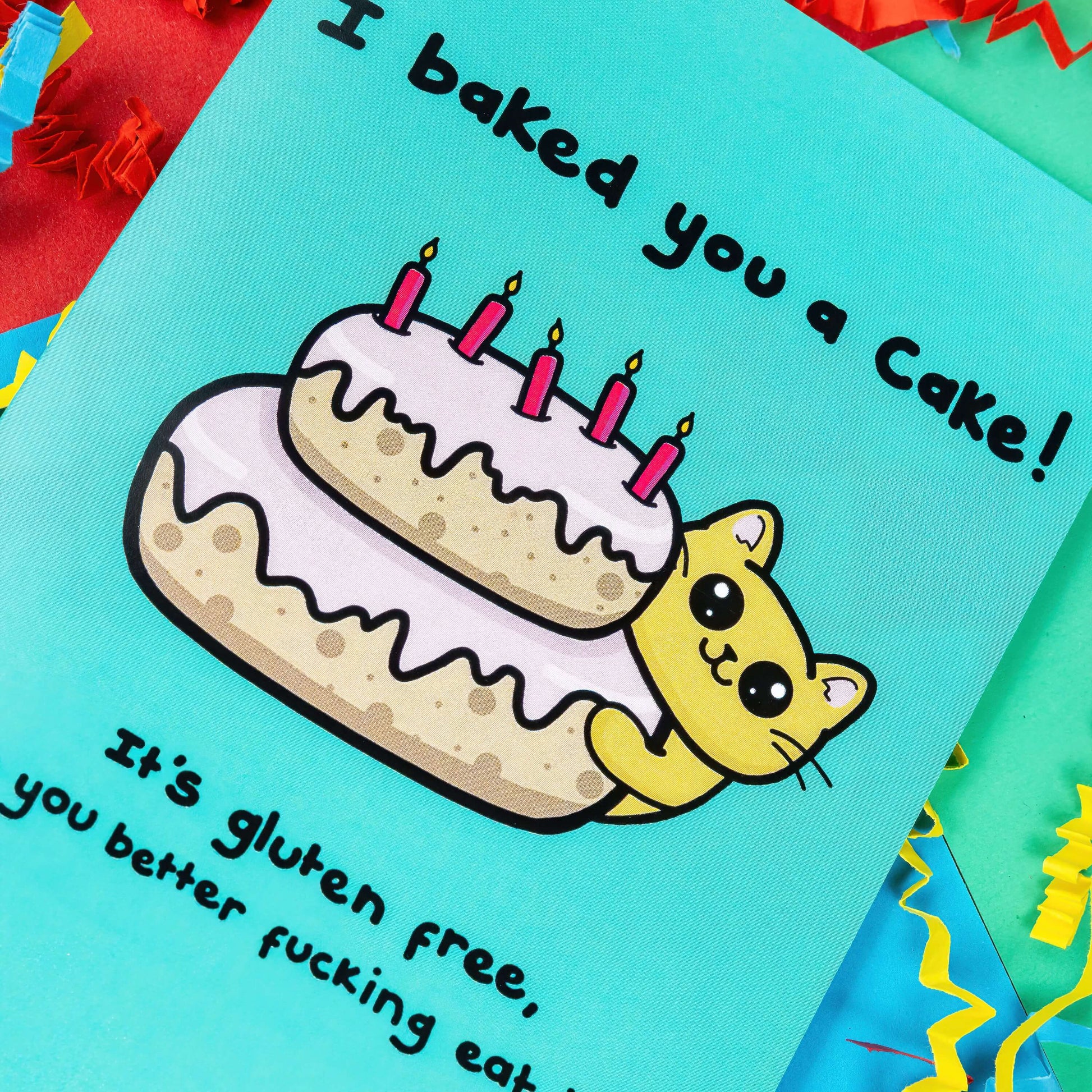A turquoise blue card that has a drawing of a yellow cat poking around a pink layered cake with candles in the top. Text on the card says I baked you a cake! It's gluten free, so you better fucking eat it! The a6 cat themed birthday card is on a blue, red and green background with red, yellow and blue crinkle card confetti. 