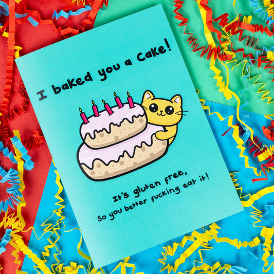 A turquoise blue card that has a drawing of a yellow cat poking around a pink layered cake with candles in the top. Text on the card says I baked you a cake! It's gluten free, so you better fucking eat it! The a6 cat themed birthday card is on a blue, red and green background with red, yellow and blue crinkle card confetti. 