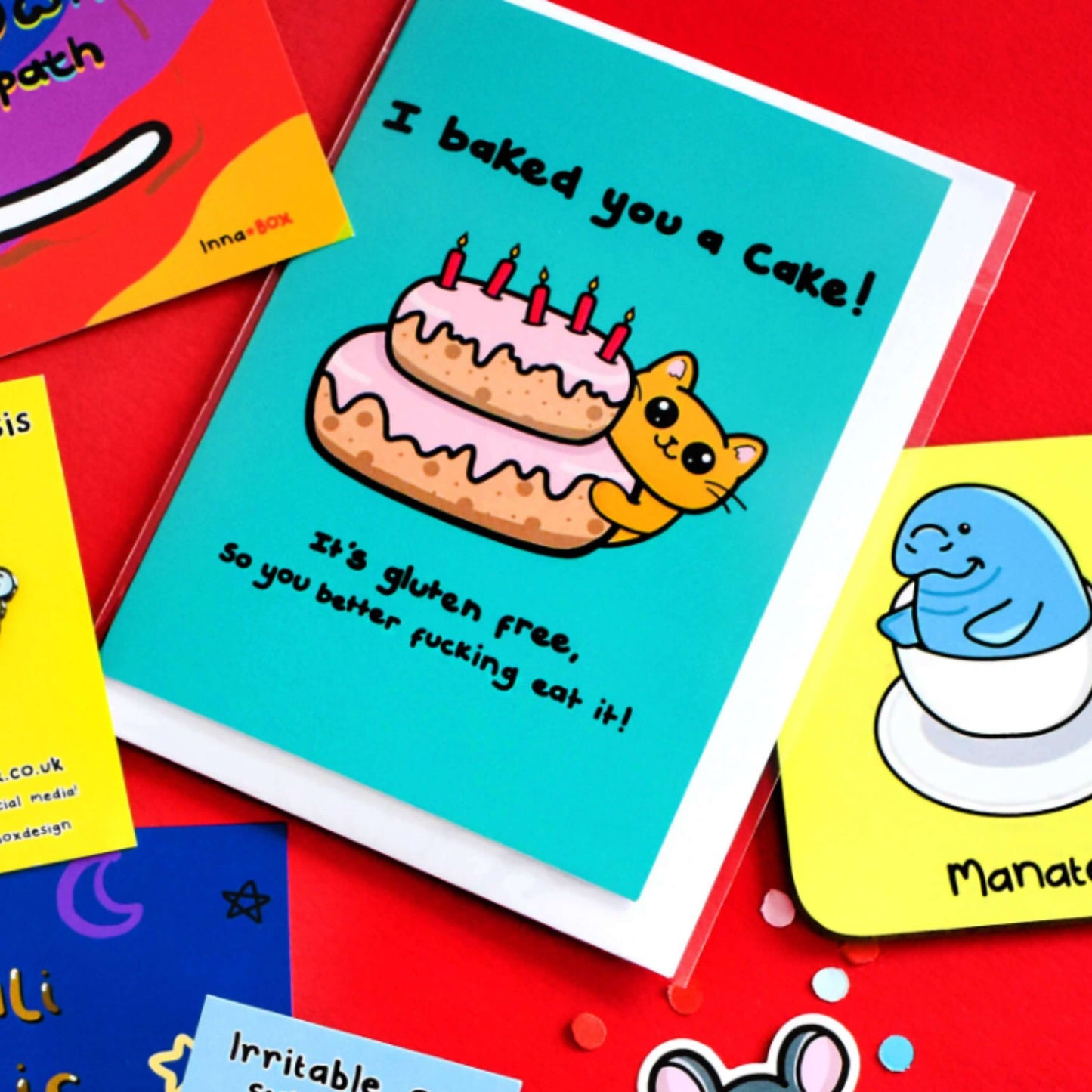 A turquoise blue card that has a drawing of a yellow cat poking around a pink layered cake with candles in the top. Text on the card says I baked you a cake! It's gluten free, so you better fucking eat it! The a6 cat themed birthday card is on a red background with other innabox products.
