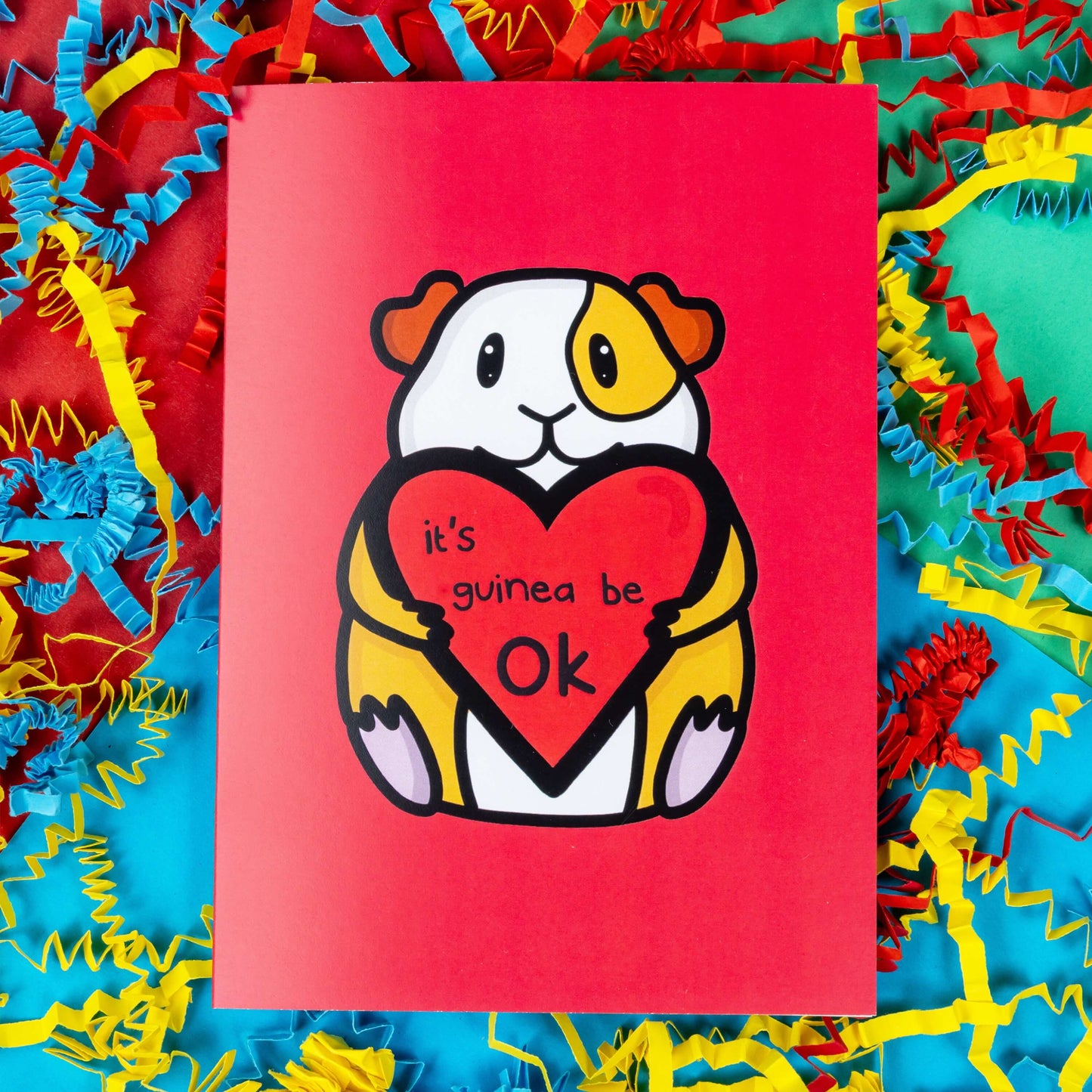 The It's Gonna Be OK Guinea Pig Card on a red, blue and green background with yellow, blue and red crinkle card confetti. The red a6 greeting card features a smiling orange and white guinea pig sat down holding a red heart with black text reading 'it's guinea be ok'. The hand drawn design is a perfect send a hug card as a gentle positive reminder.