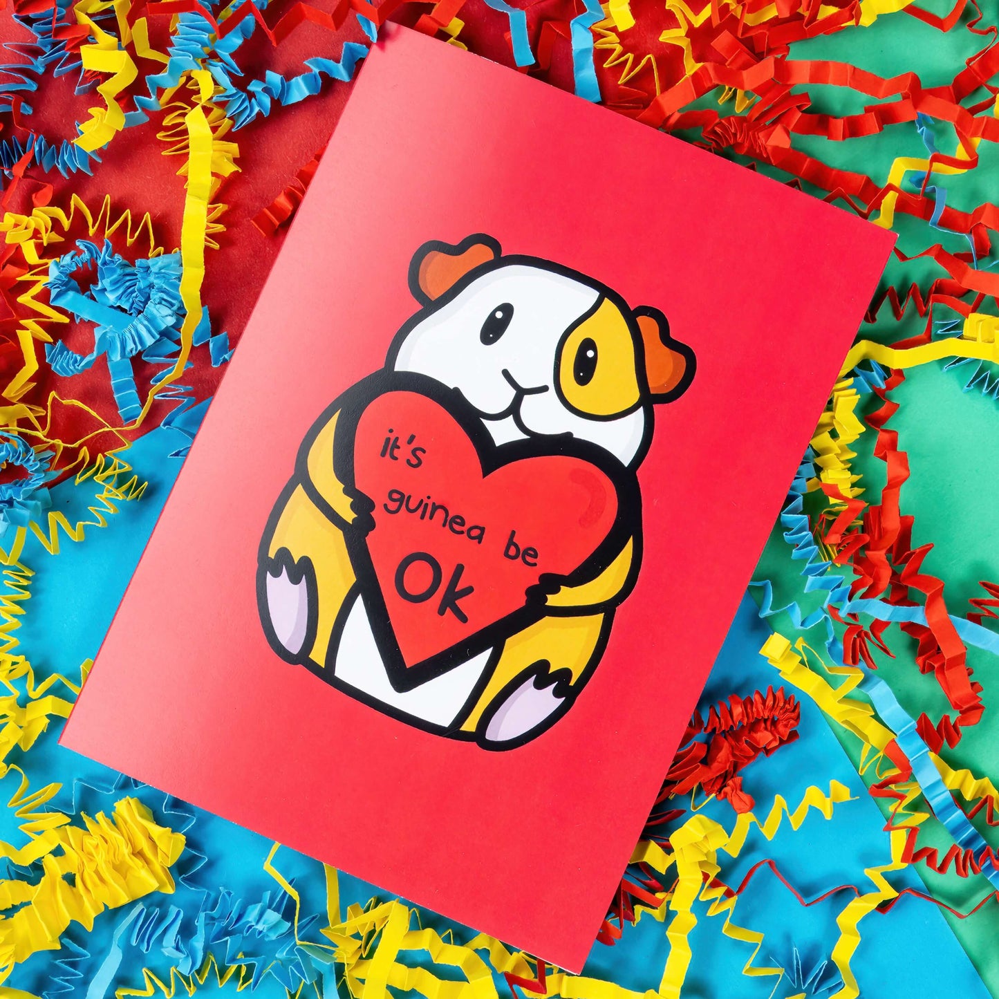 The It's Gonna Be OK Guinea Pig Card on a red, blue and green background with yellow, blue and red crinkle card confetti. The red a6 greeting card features a smiling orange and white guinea pig sat down holding a red heart with black text reading 'it's guinea be ok'. The hand drawn design is a perfect send a hug card as a gentle positive reminder.