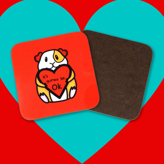 It's Guinea Be OK Coaster on a red and blue background. The red wooden coaster has a smiling guinea pig holding a big red heart with the text it's guinea be ok inside. Hand drawn design to raise awareness for mental health.