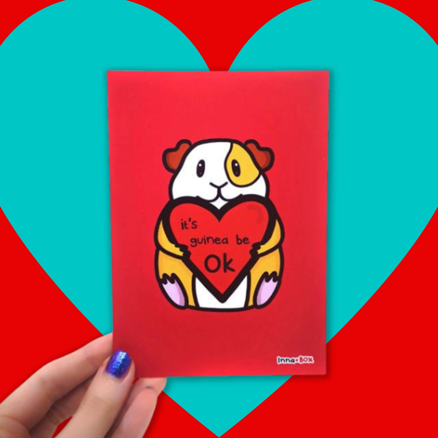 The It's Gonna Be OK Guinea Pig Postcard being held over a red and blue background. The red postcard print features a cute orange and white smiling guinea pig sat holding a red heart with black text reading 'it's guinea be ok'. The hand drawn design is a reminder for staying positive.