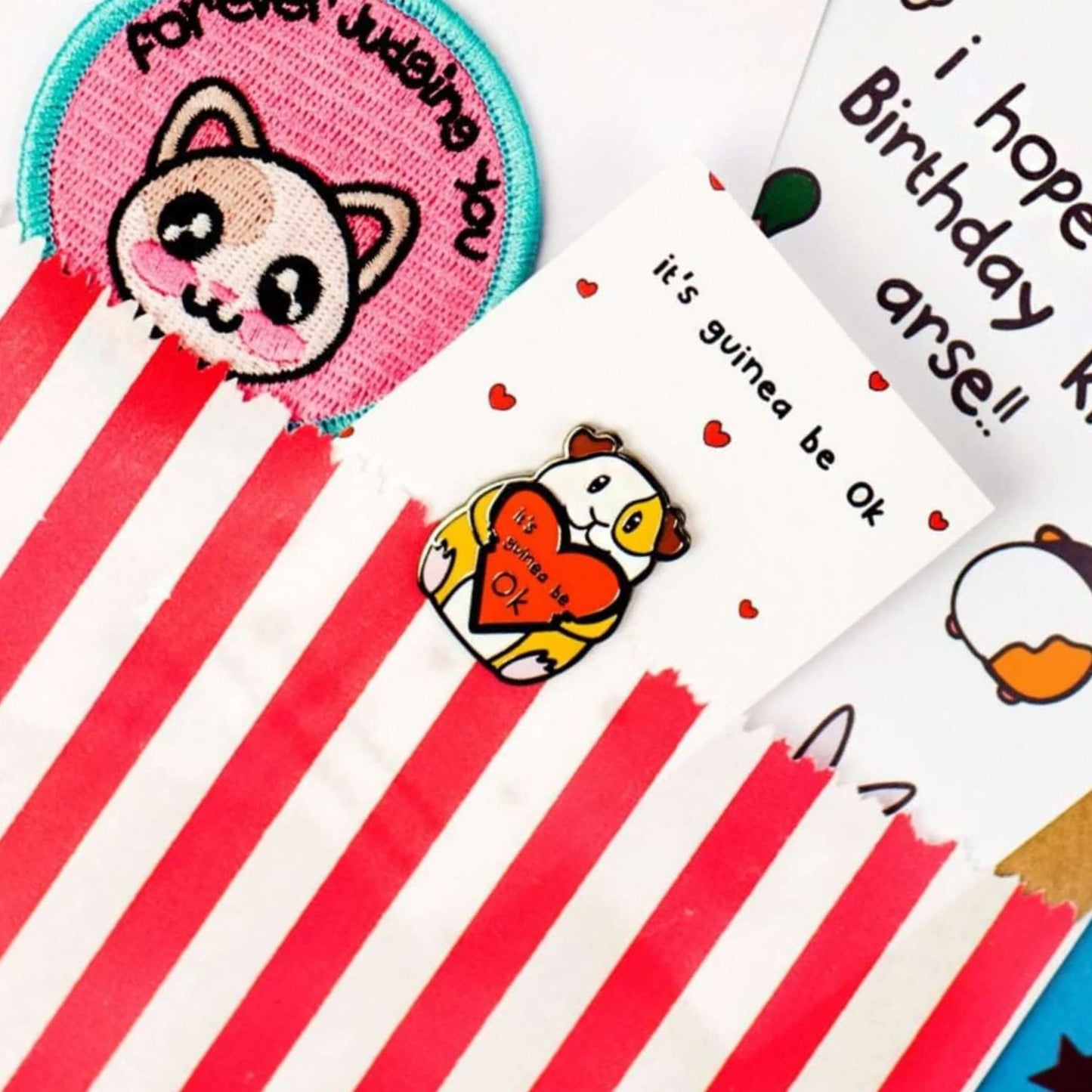 The It's Guinea Be Ok Enamel Pin on white backing card with tiny red hearts popping out of a red stripe candy bag with other innabox products.