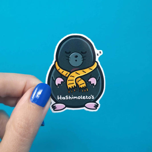A sticker of a small grey mole looking sleepy wearing a yellow scarf it has text saying hashimoletos on its lower body. It is being held over a blue background for size reference. The hand drawn design is raising awareness for Hashimotos.