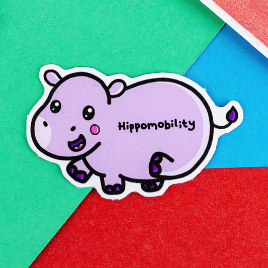 Hippomobility Sticker - Hyper Mobility on a red, blue and green background. A cute smiling pastel purple hippo flicking its back leg up with black text reading 'hippomobility' across its body. The hand drawn design is raising awareness for hyper mobility and EDS.