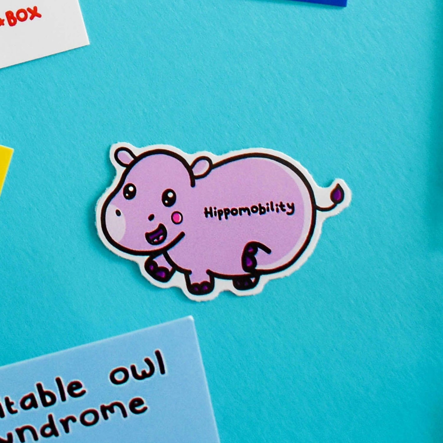 Hippomobility Sticker - Hyper Mobility on a blue background with other innabox products. A cute smiling pastel purple hippo flicking its back leg up with black text reading 'hippomobility' across its body. The hand drawn design is raising awareness for hyper mobility and EDS.